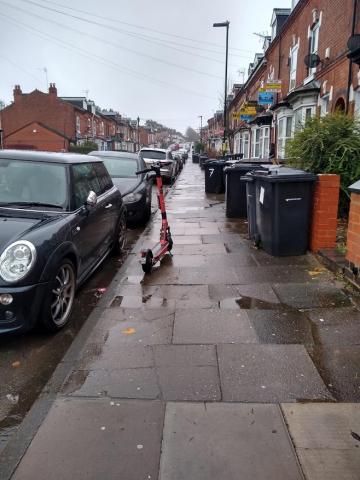 A Voi scooter parked on the pavement, narrowing the gap between a parked car and some bins